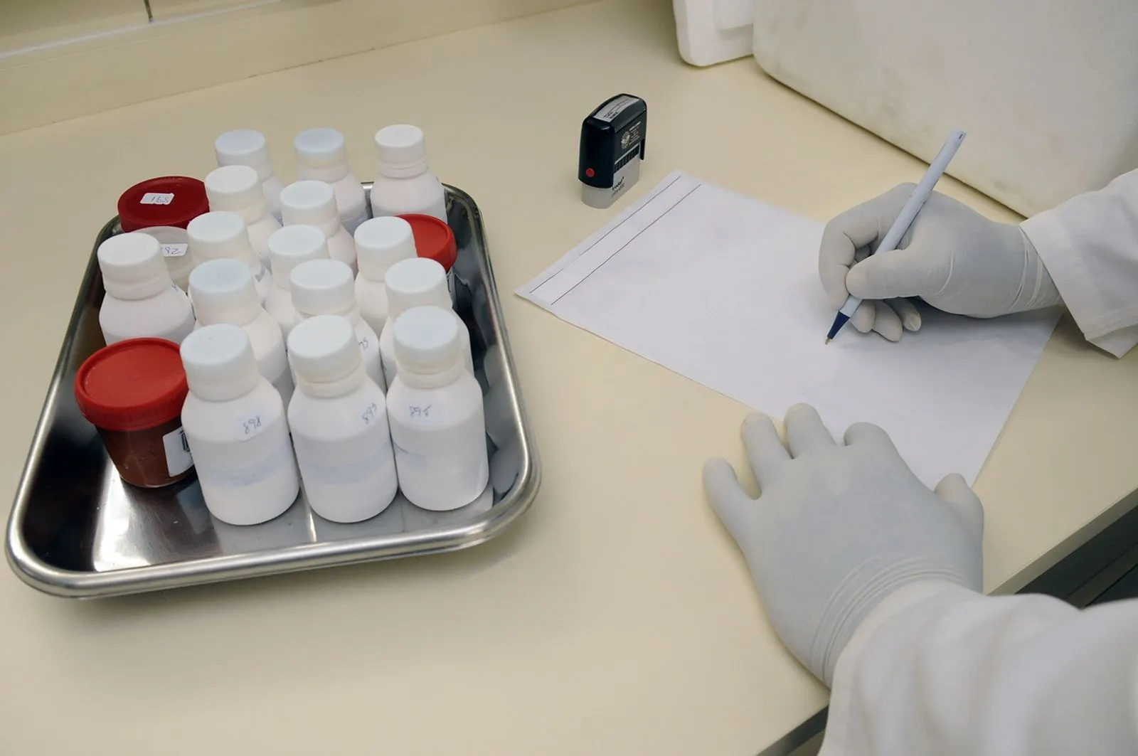 Gloved hands recording results beside a tray of labeled pharmaceutical sample bottles in a regulated laboratory.
